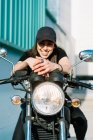 Femme motocycliste positive assise sur une moto garée dans la rue de la ville par une journée ensoleillée et regardant la caméra — Photo de stock