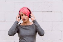 Young female with bright pink hair listening to music with headphones while standing near white wall with eyes closed — Stock Photo