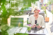 Альтернативна жінка з коротким волоссям, що переглядає соціальні медіа на смартфоні, сидячи за столом у вуличному кафе в сонячний день — стокове фото