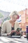 Mujer alternativa con pelo corto navegando por las redes sociales en el teléfono inteligente mientras está sentada en la mesa en la cafetería de la calle en un día soleado - foto de stock