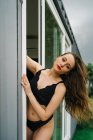 Teneur femelle mince en lingerie noire debout près de la porte en verre menant au balcon et regardant la caméra — Photo de stock