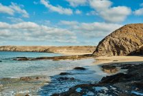 Широкоугольный вид на мирную лагуну с чистой морской водой рядом с песчаным пляжем и скалами на фоне пасмурного солнечного неба в Фуэртевентуре, Испания — стоковое фото