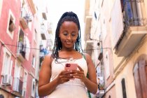 Bajo ángulo de sonriente mujer afroamericana con brads mensajería en las redes sociales a través del teléfono móvil mientras está de pie en la calle con edificios antiguos en Barcelona - foto de stock