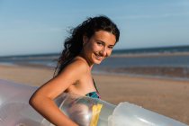 Side view of positive female in summer outfit and with inflatable mattress walking on sandy coast on sunny day during vacation — Stock Photo