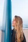 Vista lateral do surfista feminino de pé com placa SUP azul na praia de areia no verão e olhando para longe — Fotografia de Stock