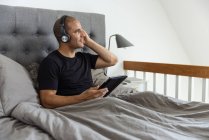 Спокойный мужчина в наушниках сидит на кровати под одеялом и просматривает социальные сети на планшете, слушая музыку по утрам — стоковое фото