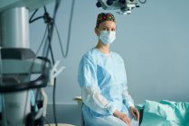 Erwachsene Ärztin in Chirurgenuniform und steriler Maske blickt in die Kamera, während sie in der Klinik sitzt — Stockfoto