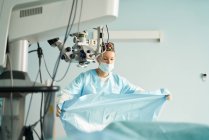 Aufmerksame erwachsene Ärztin in Einmalmaske und Zierkappe arbeitet in Klinik mit Operationsmikroskop — Stockfoto
