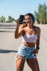 Положительная женщина-фигуристка в роликах и наушниках делает самоснимок на мобильном телефоне в солнечный день летом в городе — стоковое фото