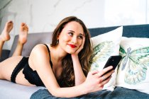 Mulher encantada em roupa interior preta deitada no sofá e mensagens nas mídias sociais via telefone celular no aconchegante lounge em casa — Fotografia de Stock
