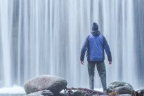 Vista posteriore di viaggiare maschio in abiti caldi in piedi sul masso e ammirando vista della rapida cascata in lunga esposizione al fiume Lozoya nel Parco Nazionale di Guadarrama — Foto stock
