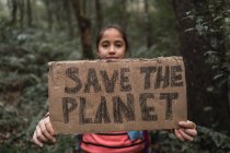 Ethnic kid raising cardboard piece with Save The Planet inscription while looking at camera in green forest — Stock Photo