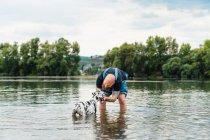 Happy man with beard in casual clothes playing with puppy while standing in lake water in summer — Stock Photo