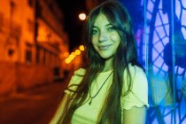 Giovane donna gentile in ciondolo e orecchino con i capelli lunghi guardando la fotocamera mentre lei è illuminata con luci verdi al neon in serata — Foto stock