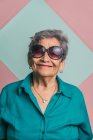 Heureuse femme âgée moderne aux cheveux gris et aux lunettes de soleil tendance sur fond rose et bleu en studio et en regardant la caméra — Photo de stock
