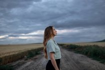 Vue latérale d'une jeune femme consciente regardant loin sur la route près d'une prairie sous un ciel nuageux en soirée à la campagne — Photo de stock