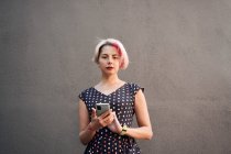 Delighted informal female with short hair and in dress messaging on mobile phone while standing against gray wall in street and looking at camera — Stock Photo