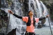 Verträumter männlicher Wanderer mit Wanderstöcken in ausgestreckten Händen, der mit geschlossenen Augen in der Nähe eines Wasserfalls im Wald steht und die Freiheit genießt — Stockfoto