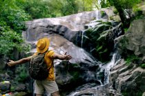 Боковой вид на неузнаваемого мужчину-туриста, стоящего на валуне и любующегося водопадом в лесу с распростертыми объятиями — стоковое фото