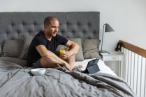 Side view of delighted male sitting on bed with glass of orange juice and browsing tablet while having breakfast in morning at home — Stock Photo