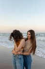 Young girlfriends embracing each other while standing on sandy beach near waving sea at sundown — Stock Photo