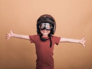 Fröhliches Kind mit Helm und Schutzbrille blickt mit ausgestreckten Armen auf beigem Hintergrund in die Kamera — Stockfoto