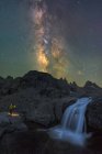 Anonymer Reisender mit Fackel betrachtet Wasserfall, der durch unwegsames felsiges Gelände unter nächtlichem Sternenhimmel mit hell leuchtender Milchstraße fließt — Stockfoto