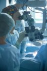 Eine erwachsene Ärztin mit steriler Maske und Zierkappe blickt durch das Operationsmikroskop gegen eine Erntehelferin im Krankenhaus — Stockfoto