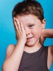 Застенчивый мальчик-подросток закрывает глаза рукой и смотрит на камеру на ярком синем фоне в студии — стоковое фото