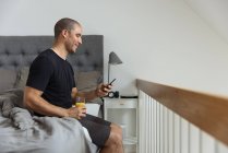 Вид збоку чоловіка сидить на ліжку, переглядаючи мобільний телефон під час сніданку вранці вдома — стокове фото