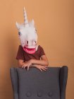 Анонимный ребенок в декоративной маске единорога с открытым ртом трогает кресло на бежевом фоне — стоковое фото
