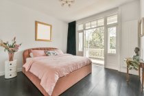 Дизайн інтер'єру будинку простора спальня з білими стінами і дерев'яною підлогою, обставлена зручним ліжком з рожевим покривалом і подушками і прикрашена квітами і макетним малюнком в денне світло — стокове фото