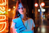 Interested young female in earrings surfing internet on mobile phone against shiny neon lamps in town — Stock Photo