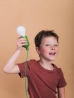 Pondering child in t shirt with plastic light bulb representing idea concept looking at camera on beige background — Stock Photo