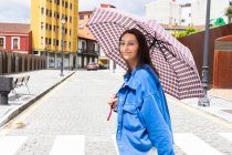 Side view of stylish female under umbrella crossing road in city and smiling — Stock Photo