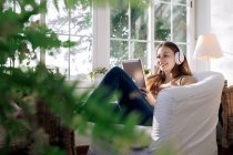 Вміст жінки в бездротовій гарнітурі, що переглядає Інтернет на планшеті, слухаючи пісню в кріслі вдома — стокове фото