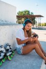 Visão lateral de positivo alegre jovem navegação feminina no telefone celular ouvir música em fones de ouvido no dia ensolarado no verão na cidade — Fotografia de Stock