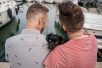 Back view of dog between cheerful bearded man embracing anonymous homosexual partner while talking and sitting on pier in harbor — Stock Photo