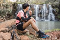 Side view of male hiker with hydration pack drinking from hose while having break during trekking in forest and admiring view of lake near waterfall — Stock Photo