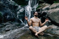Мирный мужчина без рубашки сидит в Падмасане с грязными руками и закрытыми глазами, занимаясь йогой и медитируя на мокрой скале возле водопада — стоковое фото