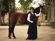 Full body of confident African American adult lady in elegant clothes and hat standing with brown horse while looking at camera near trees and building in daytime — Stock Photo