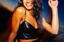 Attrayant heureux jeune brune hispanique aux cheveux longs en haut noir assis dans une voiture avec réflexion de la lumière la nuit — Photo de stock