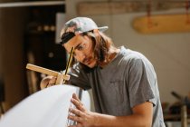 Концентрированный хипстер-мужчина с помощью инструмента писца с карандашом во время маркировки доски для сёрфинга перед формированием в мастерской — стоковое фото