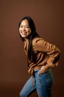 Seitenansicht einer fröhlichen Asiatin in trendiger Kleidung, die in die Kamera auf braunem Hintergrund im Studio blickt — Stockfoto
