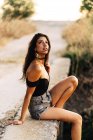 Seitenansicht der unbeschwerten barfüßigen jungen hispanischen Frau in schwarzem Top und grauer Jeanshose, die an einem Sommertag in der Natur auf einer Steinbrücke sitzt — Stockfoto