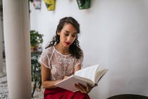 Zufriedene ethnische Frau in stylischem Outfit sitzt auf einem Hocker im Innenhof und liest Roman im Buch, während sie das Sommerwochenende genießt — Stockfoto