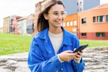 Positive young female in stylish clothes standing in city street and messaging on mobile phone while laughing with closed eyes — Stock Photo