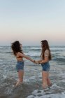 Вид збоку на молодих жінок, що тримаються за руки, стоячи в морських хвилях проти безхмарного вечірнього неба під час романтичного побачення — стокове фото