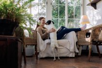 Vista lateral de alegre hembra en auriculares divirtiéndose con perro de pura raza en sillón contra ventana en casa - foto de stock