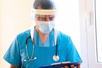 Сосредоточенный врач-мужчина со стетоскопом и в маске, читающий медицинское заключение на планшете, стоя в клинике — стоковое фото